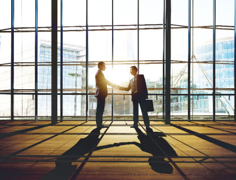 Two businessmen in front of a large window with sun streaming in shaking hands